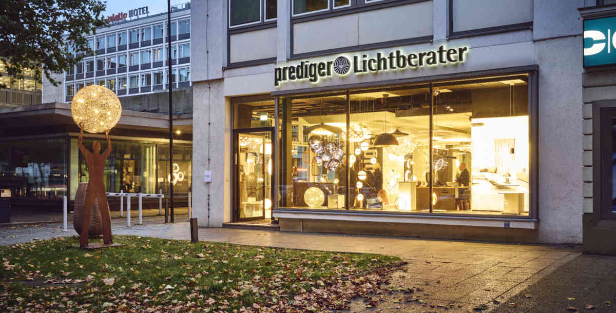 Das Bild zeigt den Showroom von Prediger Lichtberater in Berlin bei abendlicher Beleuchtung. Große Glasfronten ermöglichen einen Blick ins Innere, wo moderne Leuchten und Lichtlösungen präsentiert werden. Vor dem Gebäude steht eine Skulptur, die eine menschliche Figur darstellt, die eine leuchtende Kugel in die Luft hebt, was das Thema Licht symbolisiert.