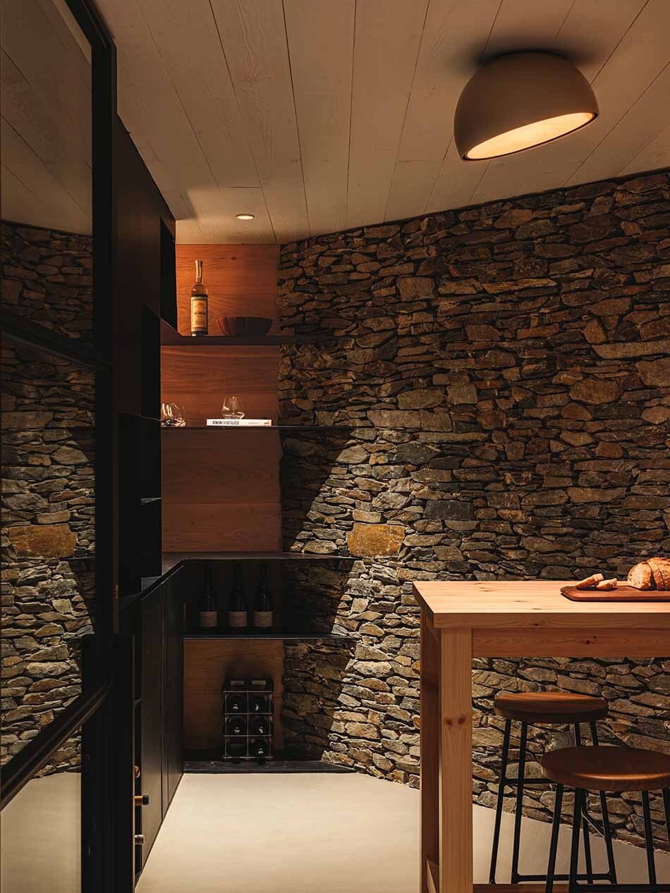 Eine gemütlicher Weinkeller mit dunkler Natursteinwand und mehreren gefüllten Weinregalen. Eine Duo Deckenleuchte von Vibia beleuchtet den Esstisch mit zwei Barhockern. Auf dem Tisch liegt geschnittenes Brot. (Foto: Vibia)