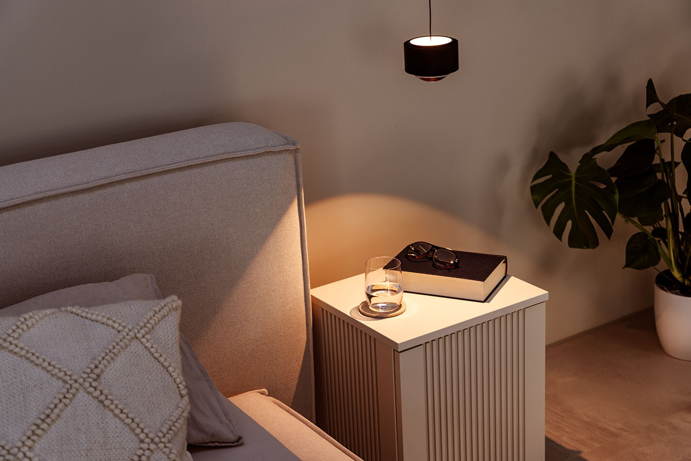 Neben einem beigem Bett hängt eine s.200 Pendelleuchte in satin schwarz von prediger.select. Die Leuchte beleuchtet einen Nachttisch, auf dem ein Wasserglas und ein Buch liegt.