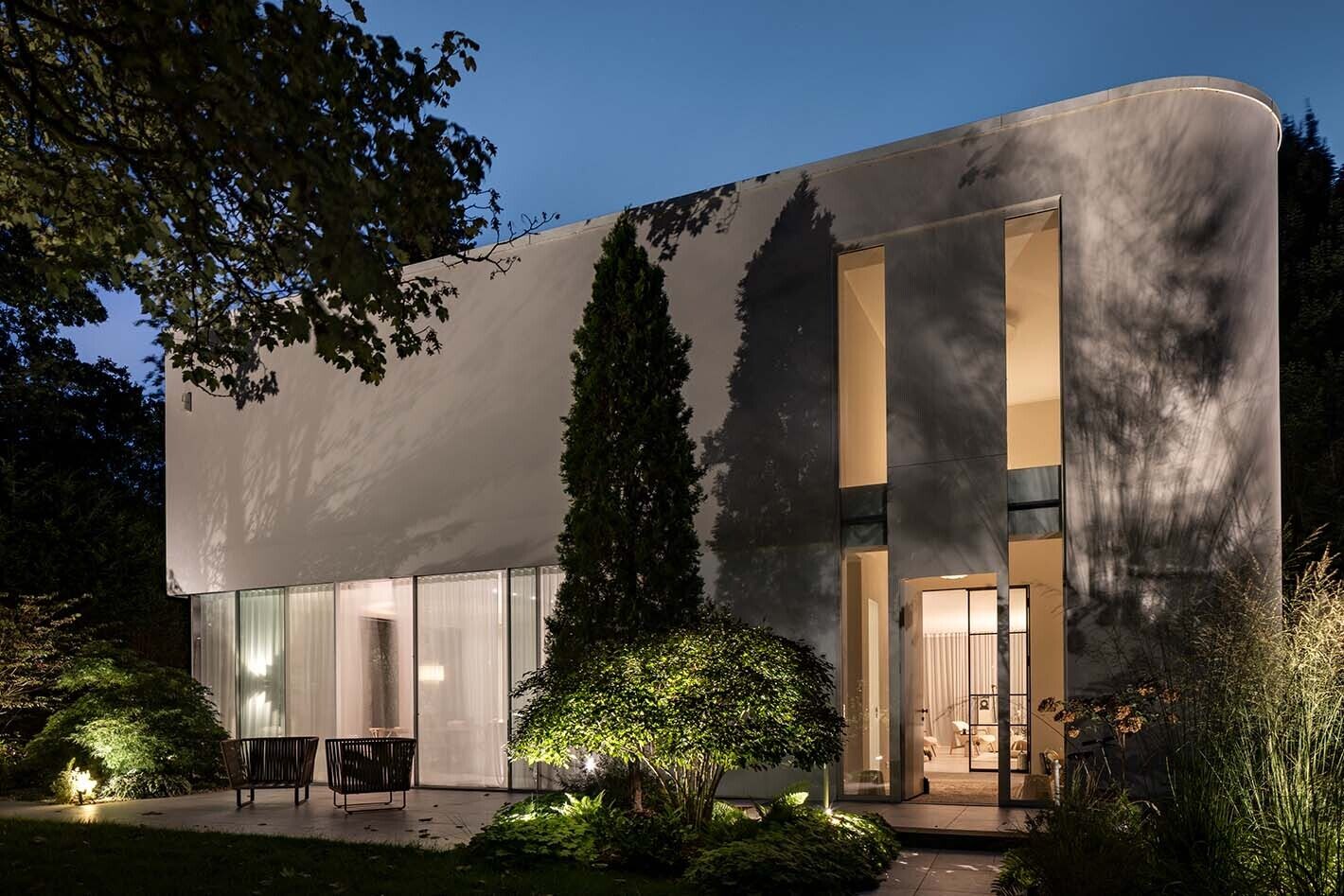Das Bild zeigt ein Haus in nächtlicher Dunkelheit, das von innen warm beleuchtet ist. Mehrere Bodenstrahler erhellen den Garten, und auch der Weg ist beleuchtet.