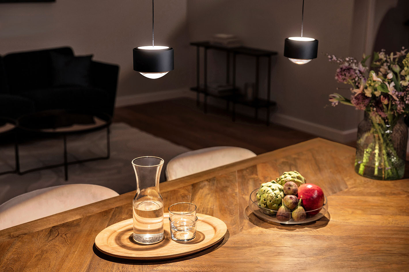 Essbereich mit einem Esstisch aus Holz, der von zwei schwarzen s.200 Pendelleuchten von prediger.select beleuchtet wird. Auf dem Tisch steht frisches Obst und Gemüse, eine Wasserkaraffe sowie ein großer Blumenstrauß.