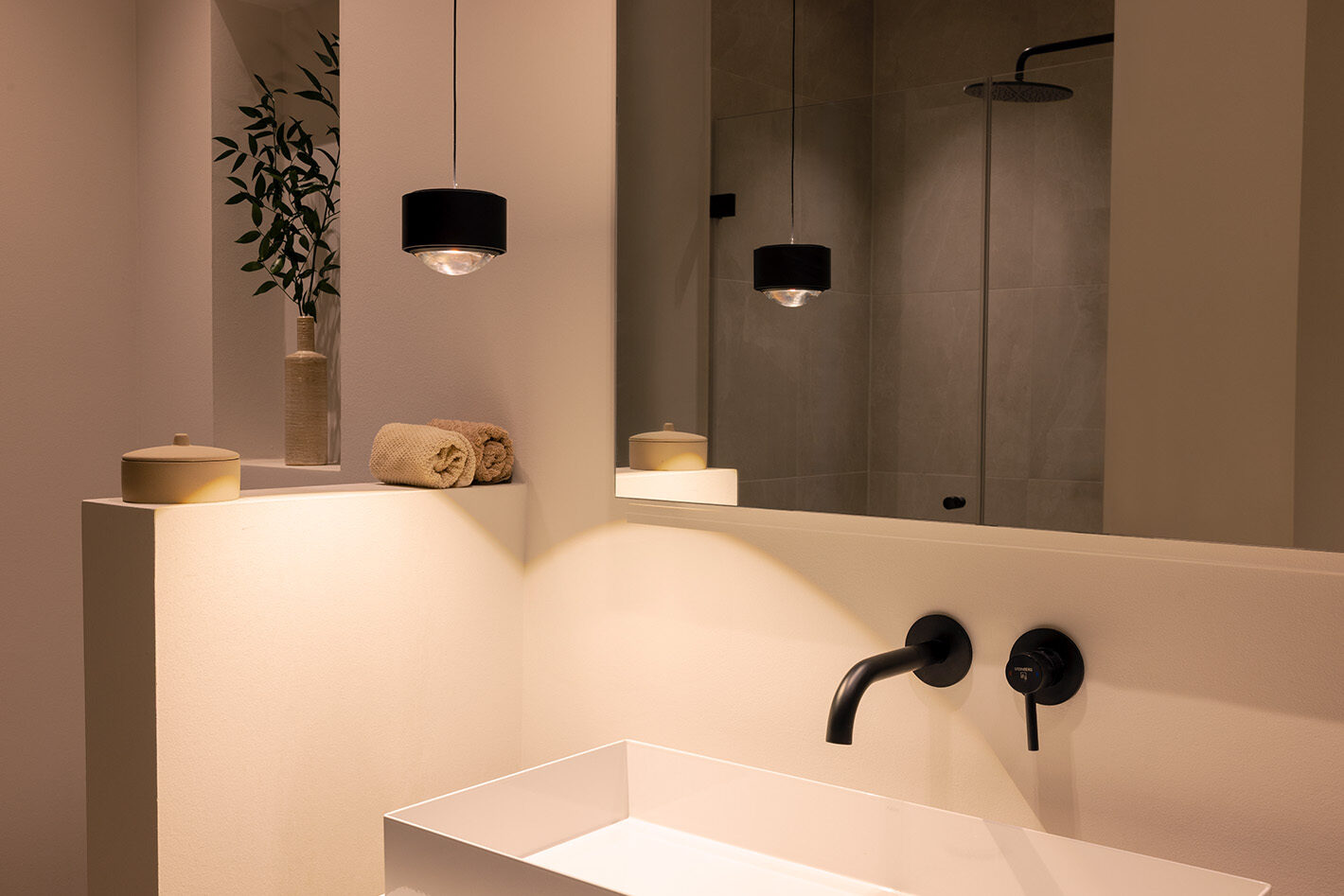 Modernes und minimalistisches Badezimmer mit einer schwarzen s.200 Pendelleuchte von prediger.select, die neben dem Waschbecken hängt. Die Leuchte strahlt Licht sowohl nach unten als auch nach oben.