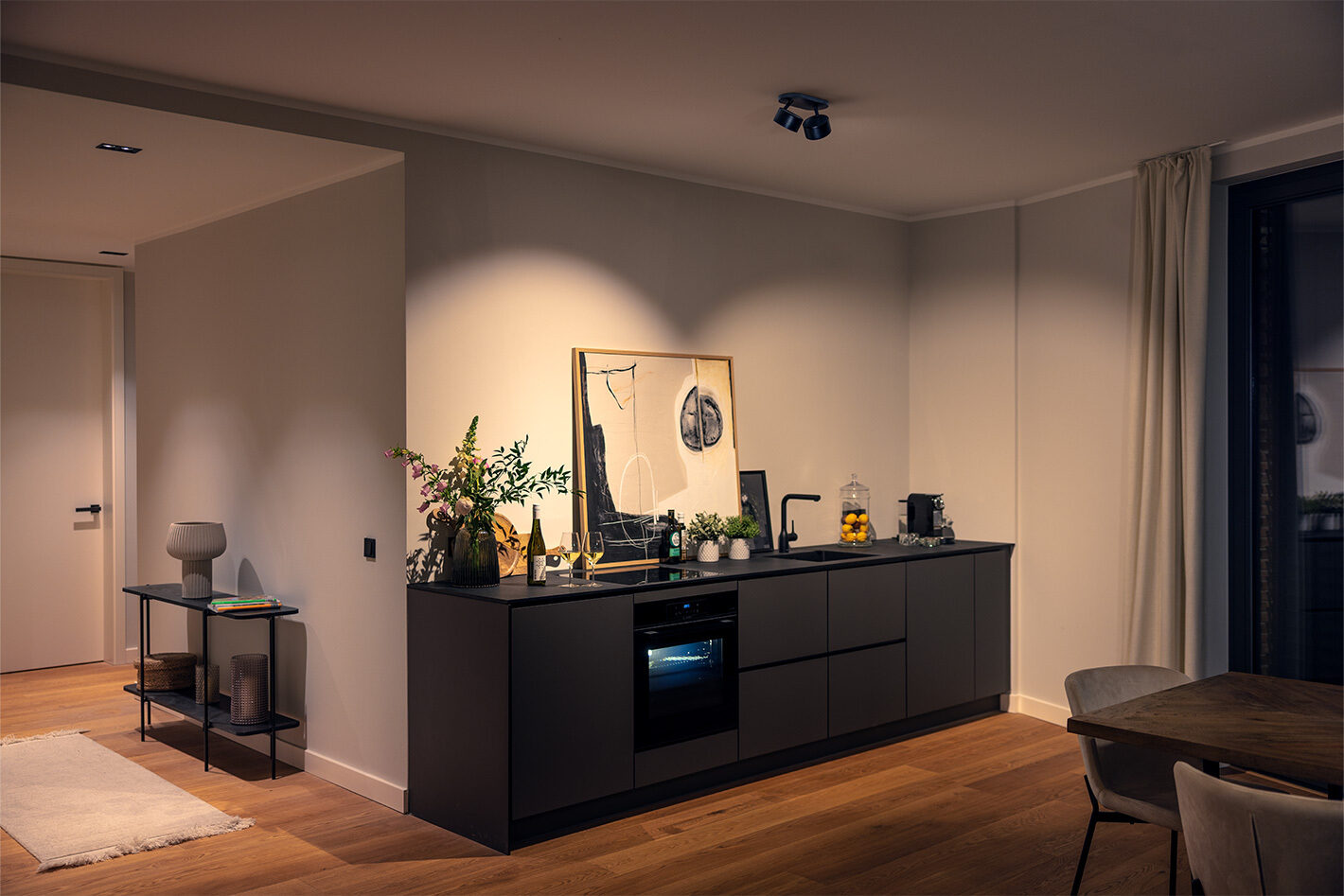 Moderner Küchenbereich mit dunkler Küchenzeile, Backofen, schwarzer Armatur, Pflanzen, Weinflaschen, abstraktem Bild an der Wand und Kaffeemaschine. Rechts Vorhang vor großem Fenster, links Beistelltisch mit Lampe und Büchern. Warme Beleuchtung durch p.011 Deckenstrahler von prediger.base, Holzboden.