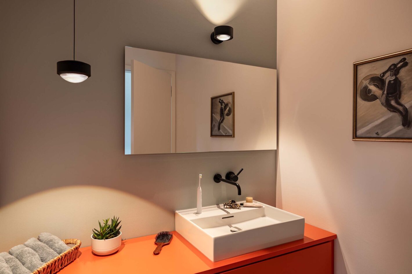 Modernes Badezimmer mit einer s.100 Pendelleuchte in Satin schwarz von prediger.select, die neben dem Waschbecken hängt. Über dem Waschbecken befindet sich ein Spiegel mit einer p.078 Wandleuchte von prediger.base. Außerdem sind zwei Zahnbürsten, eine Haarbürste, Pflanzen und Handtücher zu sehen.