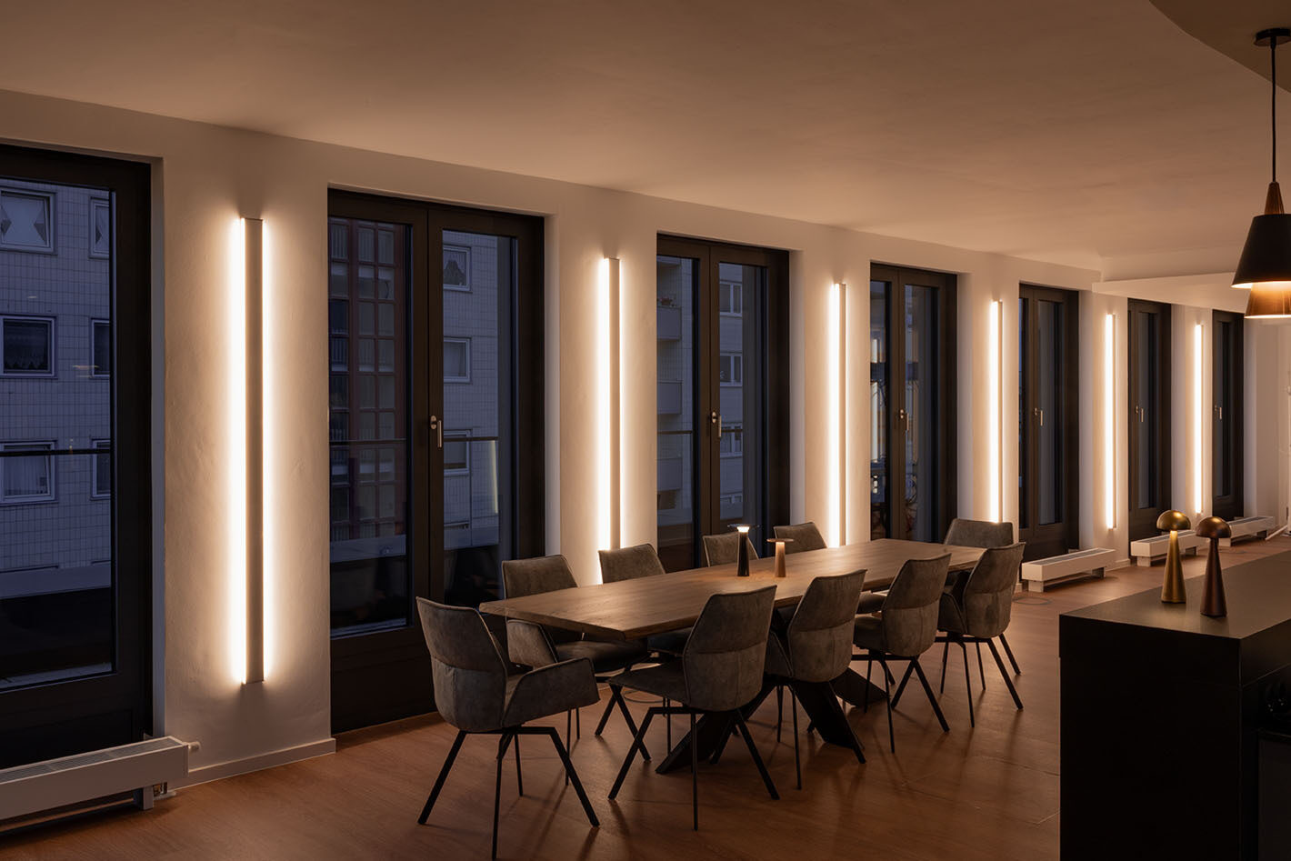 Großer Konferenzraum mit LED-Wandprofilen von Sigor und zwei Lix Akkuleuchten von IP44.DE, die auf dem langen Konfernztisch stehen. Rechts auf dem Tresen stehen zwei weitere p.141 Akkuleuchten von prediger.base.