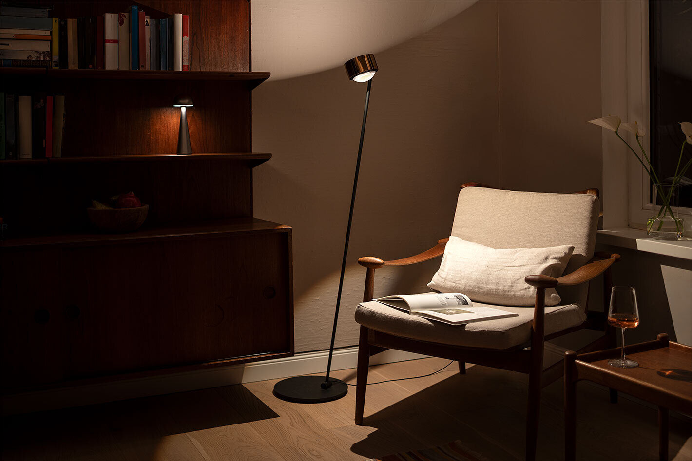 Wohnzimmer mit Bücherregal und einem skandinavischen Lesessesel aus Holz, auf dem ein aufgeschlagenes Buch liegt. Im Bücherregal steht die p.141 Akkuleuchte in Silber von prediger.base. Links neben dem Sessel steht links die s.200 Standleuchte in Gold von prediger.select und rechts befindet sich ein Beistelltisch aus Holz mit einem Weinglas.