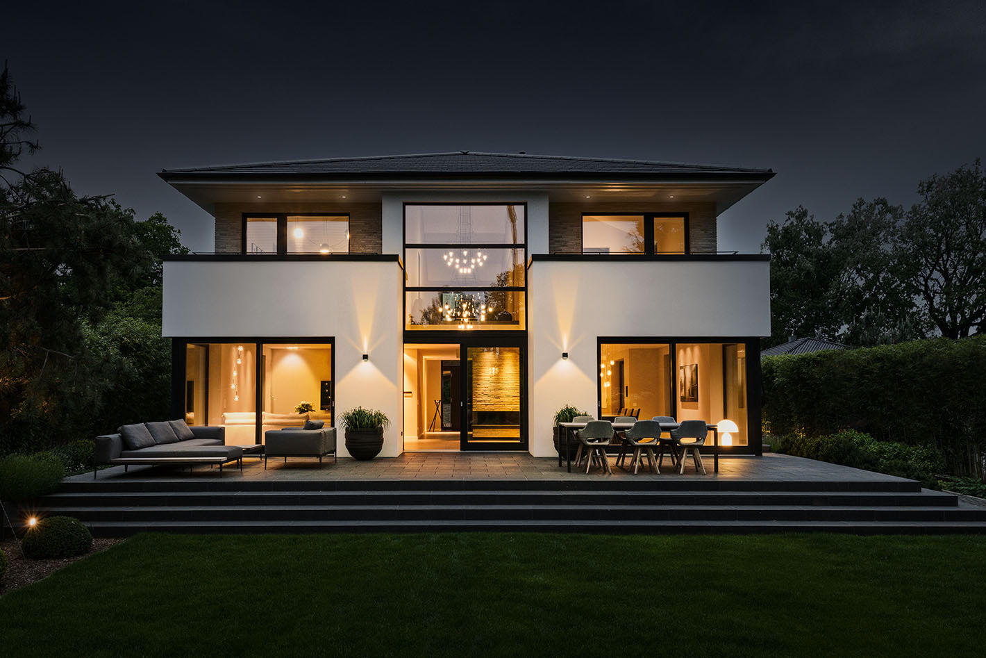 Außenaufnahme bei Abenddämmerung einer modernen Stadtvilla. Die Fassade wird von zwei p.037 Wandleuchten von prediger.base beleuchtet, die in beide Richtung strahlen. Der Innenbereich der Villa wird ebenfalls durch mehrere Lichtquellen beleuchtet.