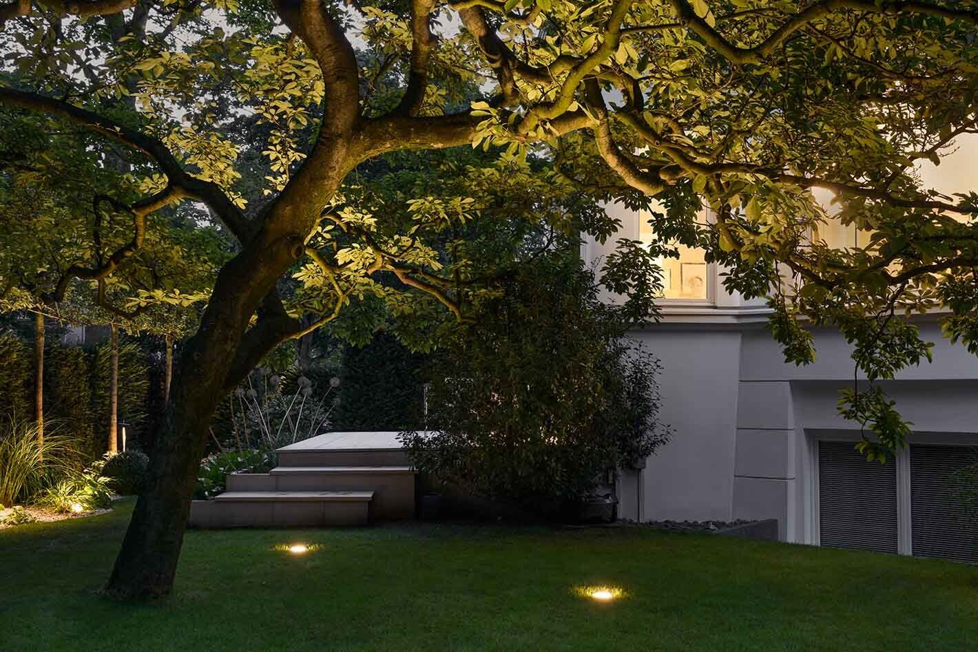 Garten mit einer Wiese und mehreren überrollbaren LED-Bodeneinbauleuchten von Bega in Edelstahl, die für Sicherheit sorgen und gleichzeitig den Baum beleuchten.