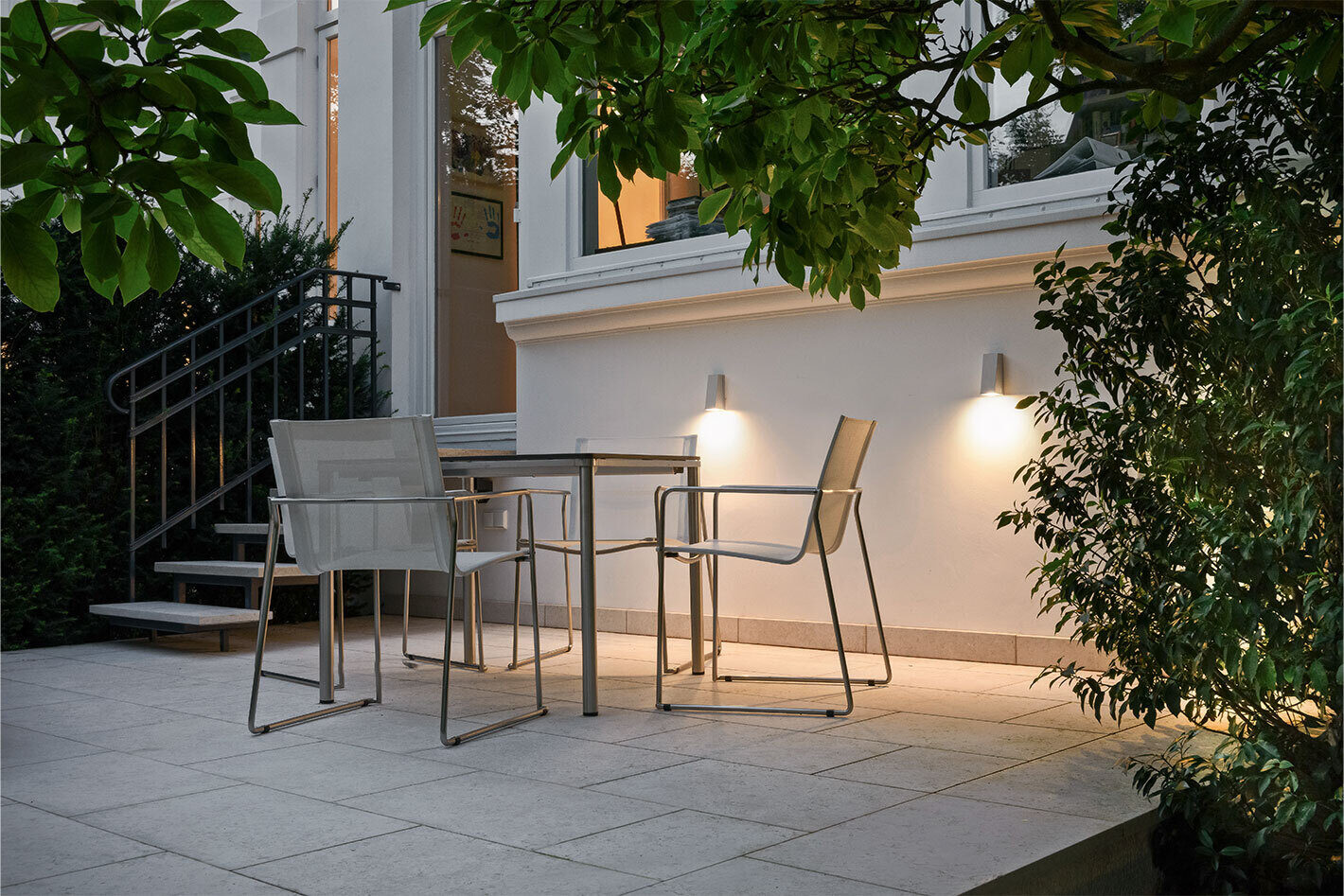 Terrasse mit zwei weißen Wandleuchten von Bega, die Licht nach unten abstrahlen. Auf der Terrasse steht ein rechteckiger Esstisch mit drei Stühlen.