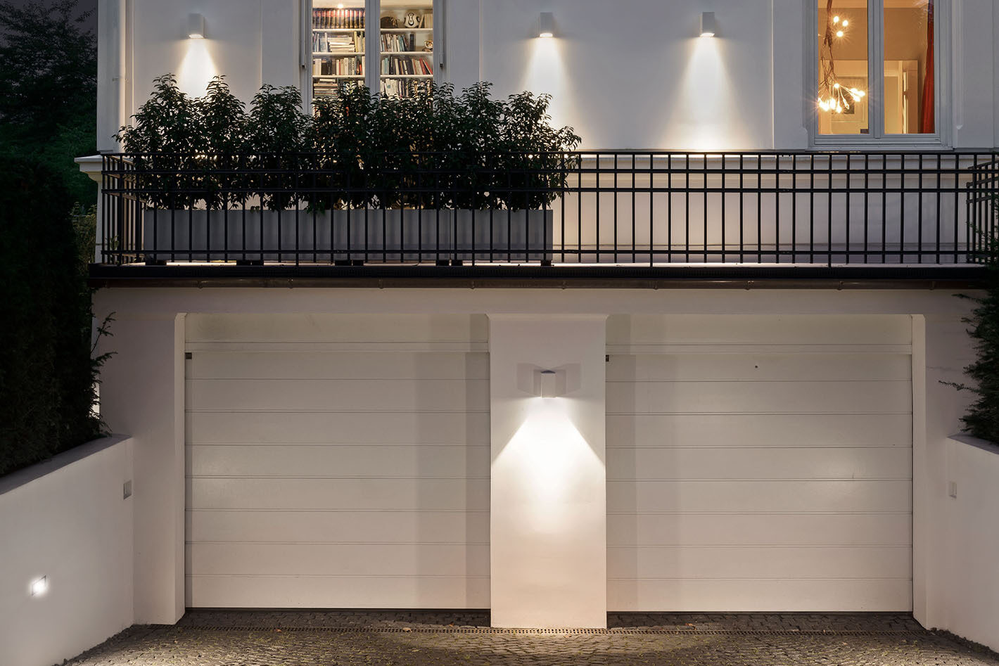 Blick auf die Garage einer Villa mit zwei Toren. Zwischen den Toren ist eine Wandleuchte von Bega montiert, die ein gezielt nach unten gerichtetes Licht abgibt. Links in der Mauer befindet sich eine dezent integrierte Einbau-Wandleuchte zur Orientierung und Sicherheit. Über der Garage eröffnet sich der Blick in den Innenbereich der Villa. An der Fassade sind mehrere Wandleuchten angebracht, die ebenfalls Licht nach unten abgeben.