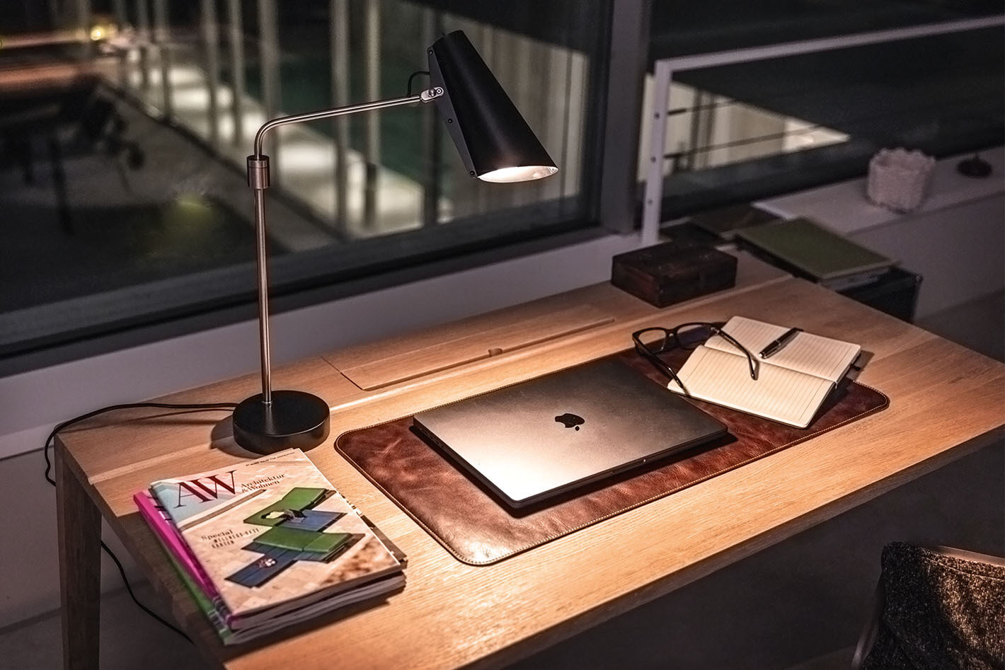 Arbeitszimmer mit einem Schreibtisch, auf dem eine Birdy Tischleuchte von Northern in schwarz steht. Sie beleuchtet ein Macbook, Magazine und ein Notizbuch.