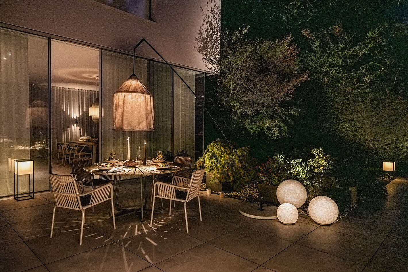 Moderne große Terrasse mit vielen unterschiedlichen Lichtquellen. Auf dem runden Esstisch steht eine Africa Akku-Tischleuchte von Vibia. Auf dem Boden stehen drei unterschiedlich große p.111 Kugelleuchten von prediger.base. Vor den bodentiefen Fenstern des Hauses steht eine Fil-Akkuleuchte von der Marke IP44.DE. Der Innenbereich wurd ebenfalls beleuchtet mit einer Plivello Pendelleuchte in grün von Le Klint sowie mit einer Stehleuchte s.200 von prediger.select in Schwarz.