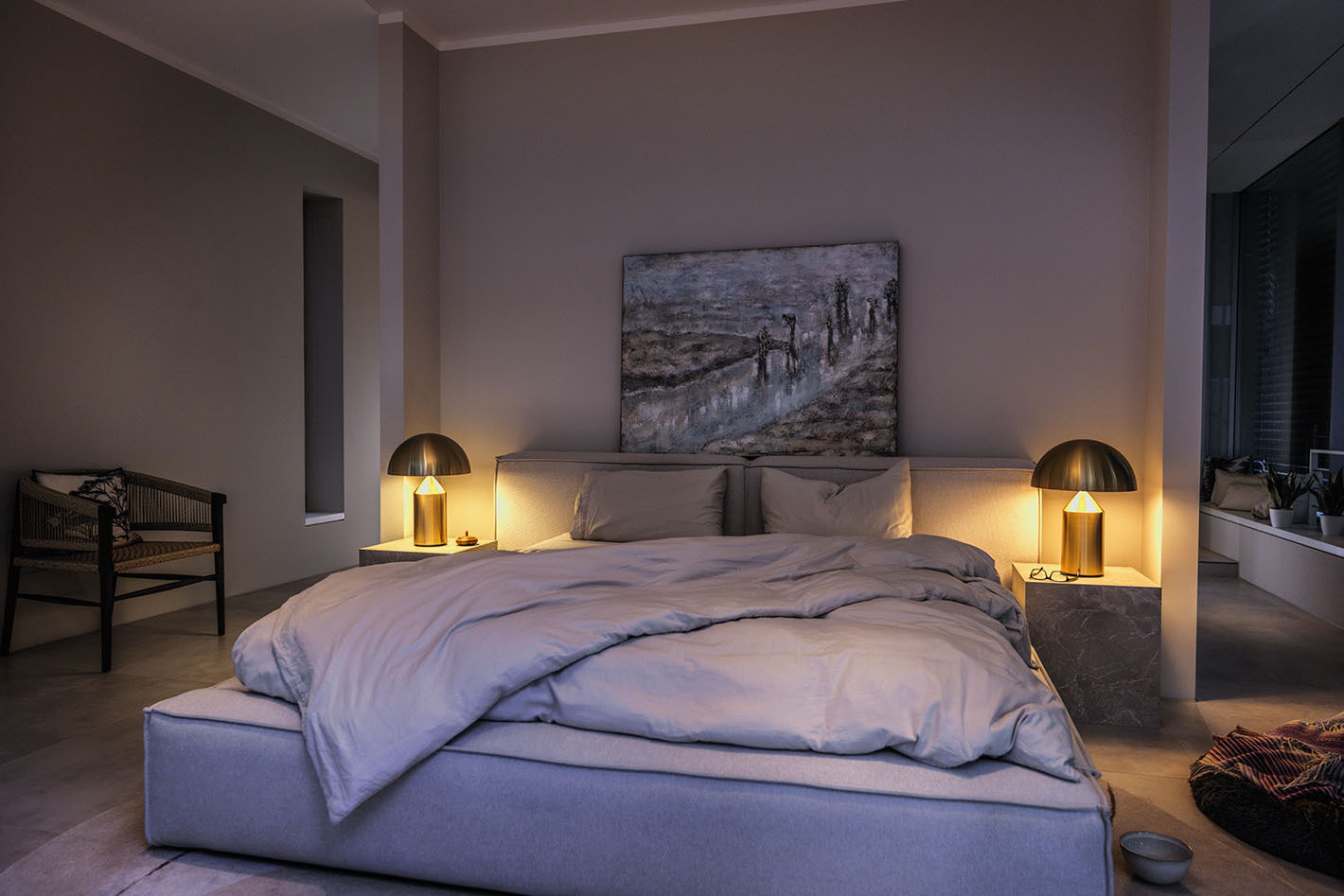 Gemütliches Schlafzimmer mit großem, grauem Bett und hellgrauer Bettdecke. Auf beiden Seiten des Bettes stehen Nachttische aus grauem Marmor, auf denen goldene Atollo Leuchten von Oluce leuchten und warmes Licht spenden. Über dem Bett hängt ein abstraktes Gemälde. Im Hintergrund links steht ein schwarzer Stuhl, rechts ist ein Fenster mit Pflanzen auf der Fensterbank. Der Raum wirkt ruhig und stilvoll eingerichtet.