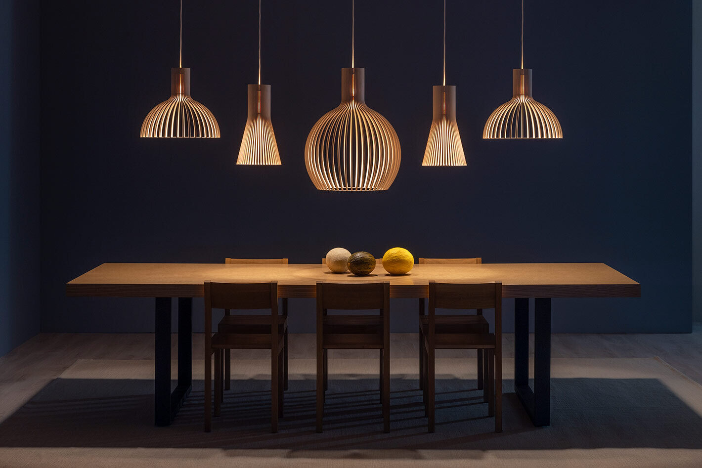 Mehrere Pendelleuchten von Secto Design hängen über einem großen Esstisch in einem modernen Wohnraum. Die Leuchten aus filigranen Holzlamellen zeigen unterschiedliche Formen und spenden warmes, nach unten gerichtetes Licht. Um den Tisch stehen mehrere Holzstühle, auf der Tischmitte liegen dekorative Kugeln. Der dunkle Hintergrund betont die skandinavische Gestaltung der Leuchtenmodelle Victo, Secto, Octo.
