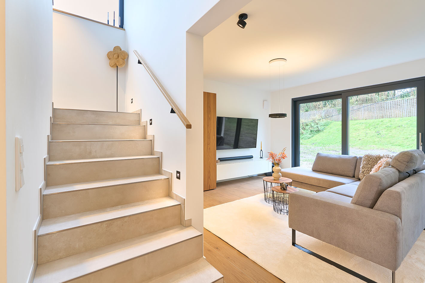 Modernes Wohnzimmer mit grauem Sofa und einer schwarzen c.Space Pendelleuchte von CHRISTOPH, die über zwei kleinen Beistelltischen hängt. Die Wohnzimmerwand wird ebenfalls durch zwei schwarze p.011 Deckenstrahler beleuchtet. Links ist die Treppe zu sehen. Die Treppenstufen werden von dreip. 076 Abgeblendete LED Wand-Einbauleuchten von prediger.base beleuchtet.