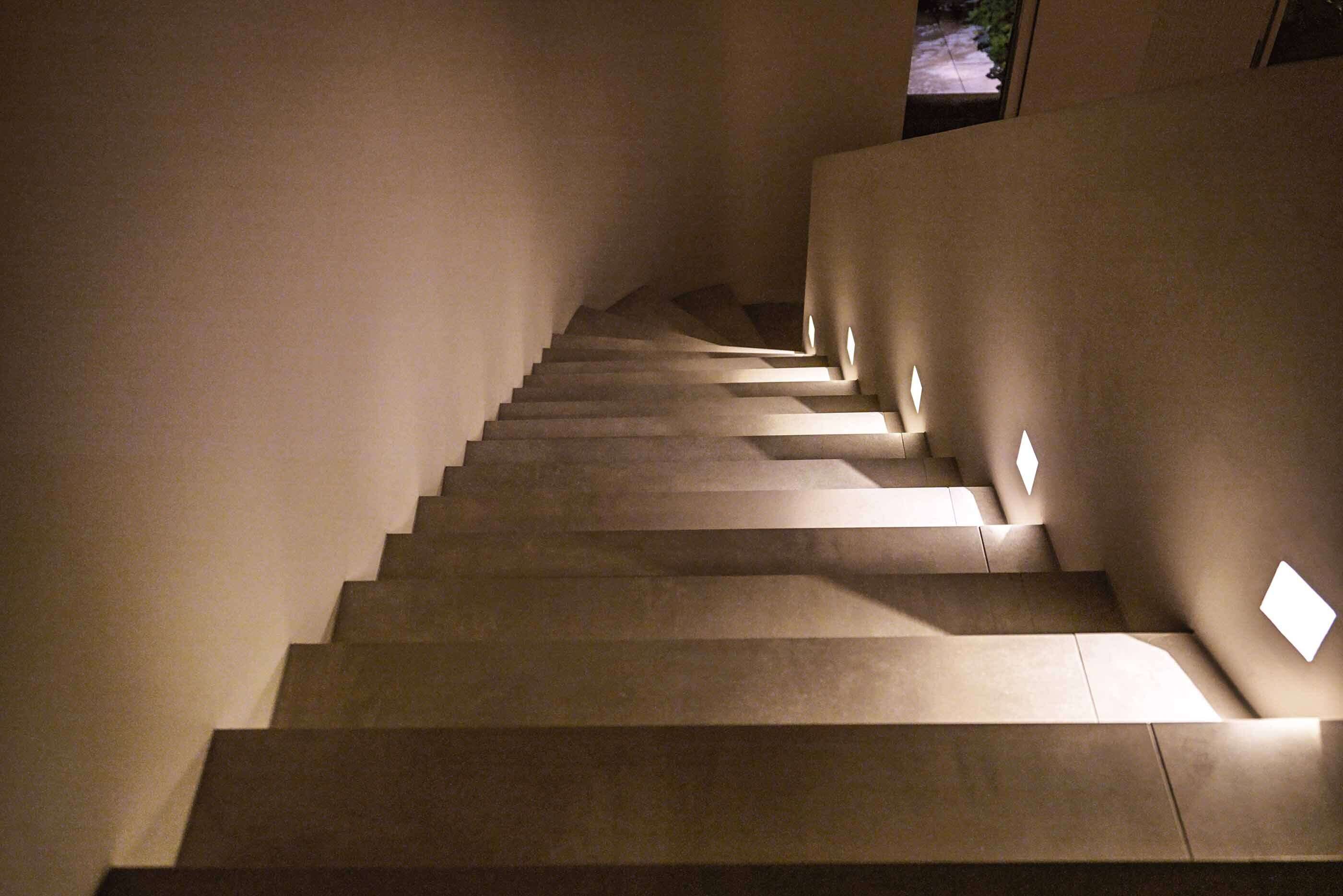 Blick von oben auf eine Treppe, die zur Haustür führt. Mehrere p.108 Wandleuchten von prediger.base beleuchten die Stufen und sorgen für Orientierung. Der Treppenbereich ist minimalistisch und schlicht.