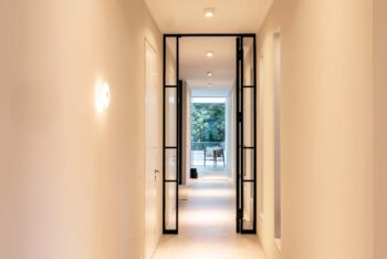 Blick in einen langen, schmalen Flur, der von mehreren  Deckenleuchten p.078 von prediger.base beleuchtet wird. Links an der Wand wurde eine Funnel Wandleuchte von Vibia montiert. Der Flur ist schlicht und minimalistisch. Die Deckenleuchten sorgen für Orientierung.
