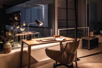 Arbeitszimmer mit Schreibtisch am Fenster, auf dem eine schwarze Birdy Tischleuchte von Northern steht. Sie beleuchtet den gesamten Tisch aus Holz. Durch die große Fensterfront blickt man nach draußen.