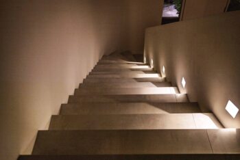 Blick von oben auf eine Treppe, die zur Haustür führt. Mehrere p.108 Wandleuchten von prediger.base beleuchten die Stufen und sorgen für Orientierung. Der Treppenbereich ist minimalistisch und schlicht.