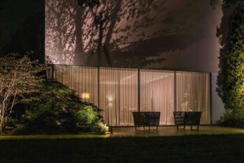 Garten während der Abenddämmerung mit p.115 Bodenstrahlern von prediger.base, die einen großen Busch beleuchten. Durch die bodentiefen Fensterfronten blickt man in den beleuchteten Innenbereich der Villa.