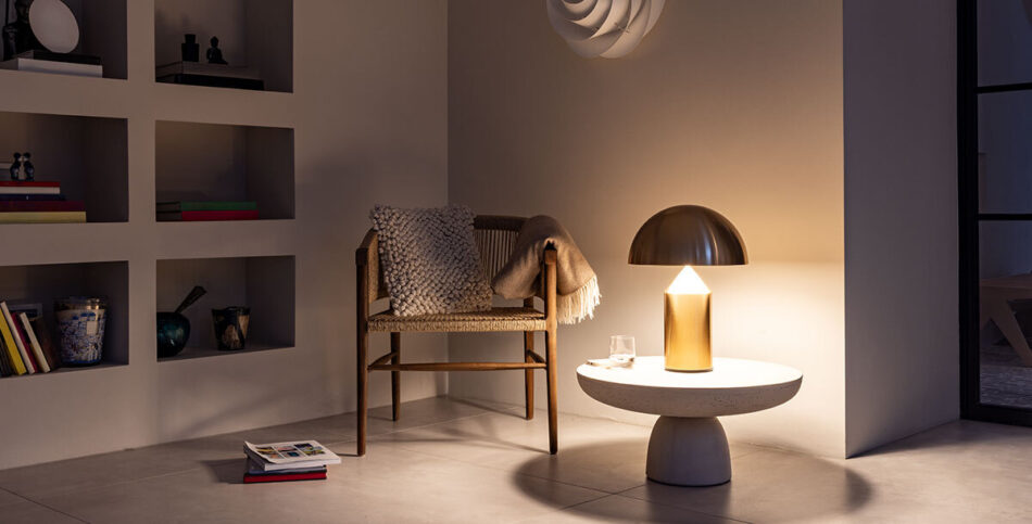 Eine gemütliche, minimalistische Wohnszene. Ein Holzstuhl mit geflochtenem Sitz und Decke steht neben einem runden Beistelltisch. Auf dem Tisch befindet sich die Tischleuchte Atollo in Gold von der Marke Oluce, die warmes Licht ausstrahlt. Im Hintergrund ist ein Einbauregal mit verschiedenen dekorativen Elementen und Büchern zu sehen. Über dem Stuhl hängt die Swirl Wandleuchte von Le Klint an der Wand.