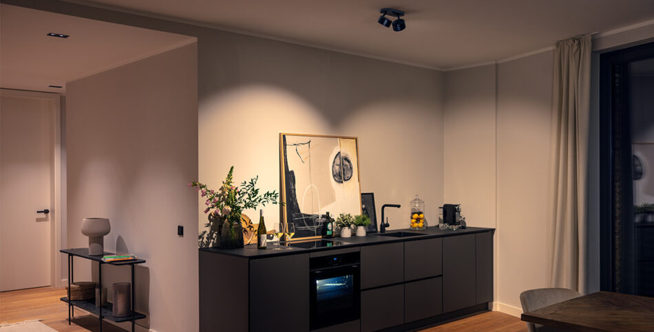 Moderner Küchenbereich mit dunkler Küchenzeile, Backofen, schwarzer Armatur, Pflanzen, Weinflaschen, abstraktem Bild an der Wand und Kaffeemaschine. Rechts Vorhang vor großem Fenster, links Beistelltisch mit Lampe und Büchern. Warme Beleuchtung durch p.011 Deckenstrahler von prediger.base, Holzboden.