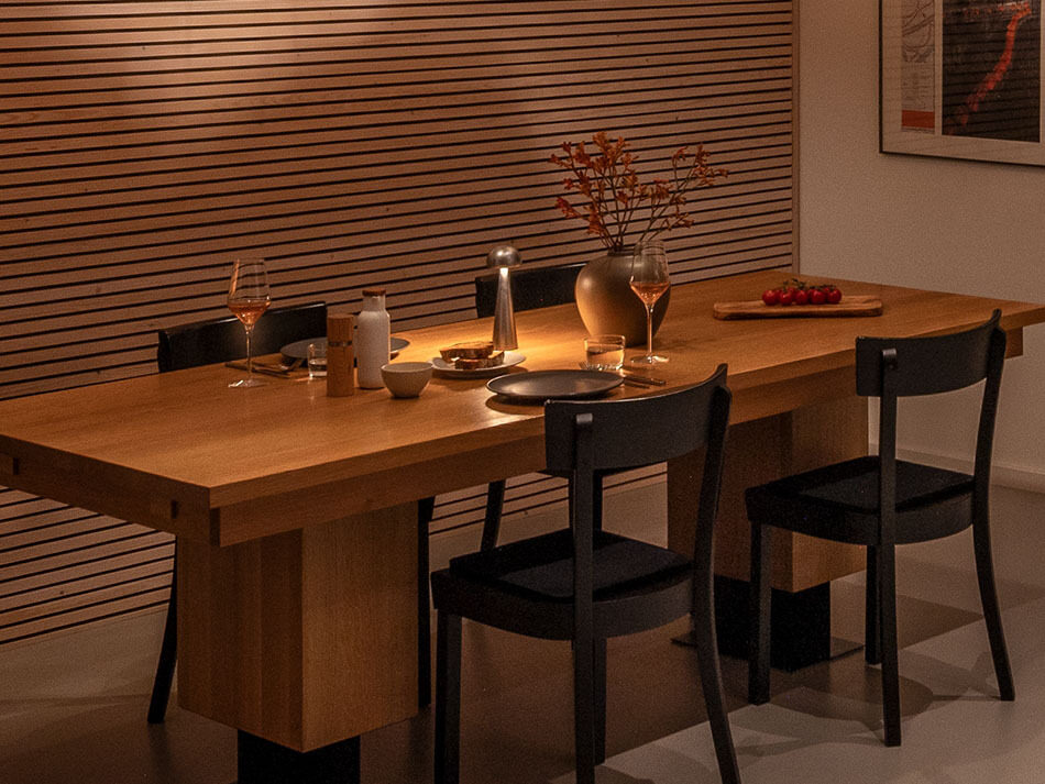 Holzesstisch mit einer p.141 Akku-Tischleuchte in Silber von prediger.base, die zusätzlich zu den Pendelleuchten Licht bietet. An dem Tisch stehen vier schwarze Stühle. Auf dem gedeckten Tisch stehen gefüllte Weingläser, Tomaten und ein Blumenstrauß.