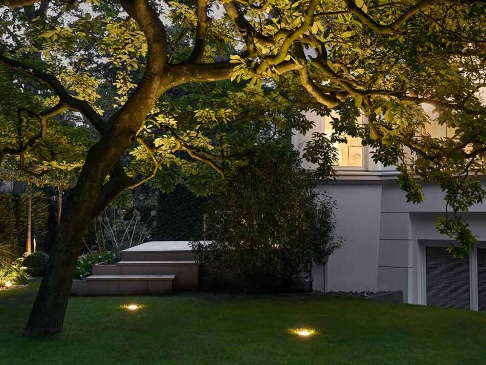 Garten mit Wiese mit Bodenleuchten von Bega, die einen Baum von unten beleuchten und für Orientierung sorgen.