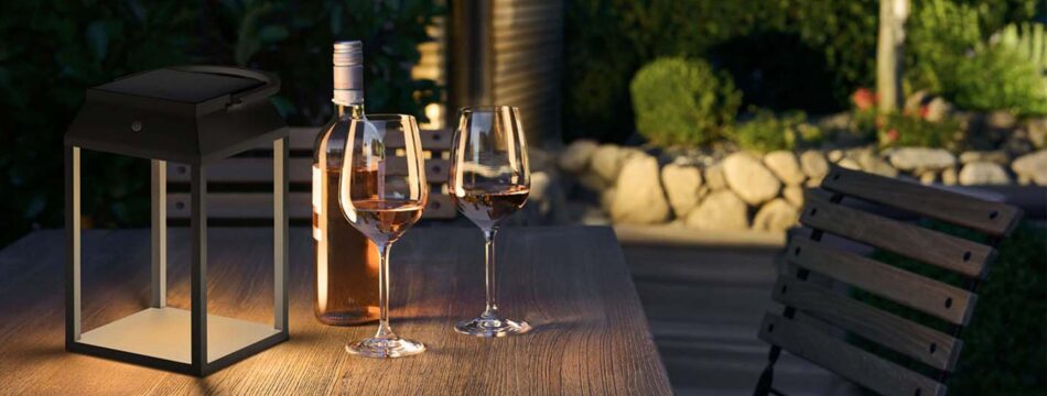 Die p.089 LED-Tischleuchte von prediger.base beleuchtet einen kleinen Tisch im Außenbereich, auf dem eine Flasche Wein und zwei halbvolle Gläser stehen. Das Solarmodul lädt den Akku der Laterne bei Sonneneinstrahlung auf. (Foto: Prediger Lichtberater)