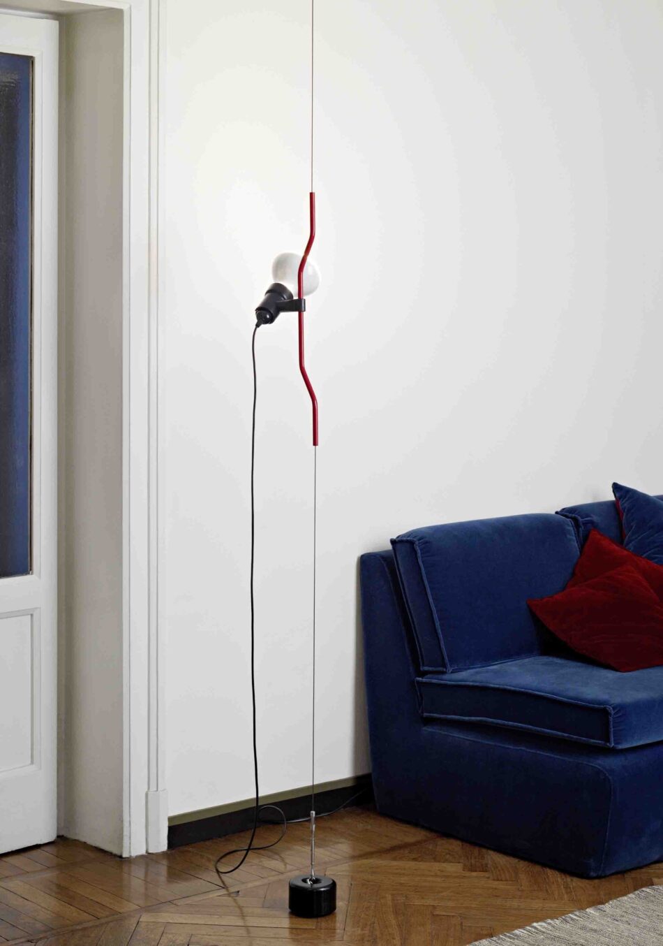 Wohnzimmer mit Parentesi Deckenleuchte von Flos, montiert an einer weißen Wand neben einem blauen Sofa mit roten Kissen. (Foto: Flos)