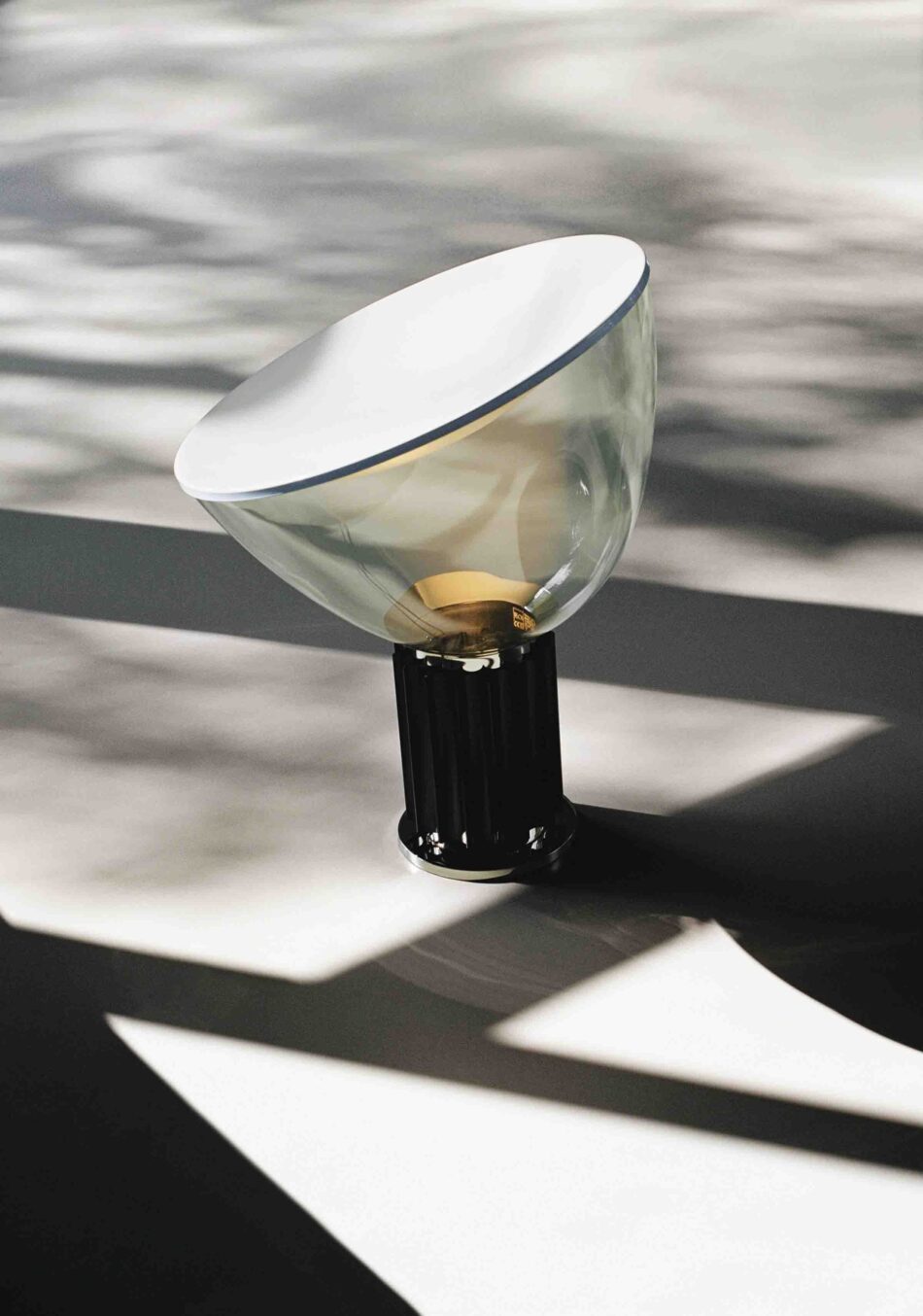 Terrasse mit Taccia Tischleuchte von Flos mit halbkugelförmigem, nach oben geöffnetem Reflektor und schwarzem geripptem Fuß. Die Leuchte steht auf einer hellen Fläche mit Schattenmustern. (Foto: Flos)