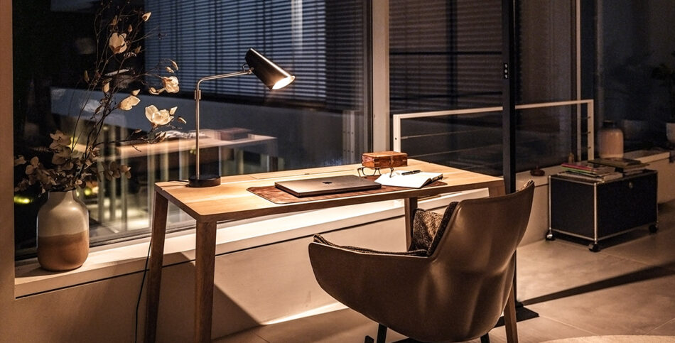 Moderner, minimalistisch eingerichteter Arbeitsbereich mit Holzschreibtisch. Darauf steht eine schwarze Birdy Tischleuchte von Northern mit silbernen Akzenten, die einen Laptop und ein Notizbuch beleuchtet.