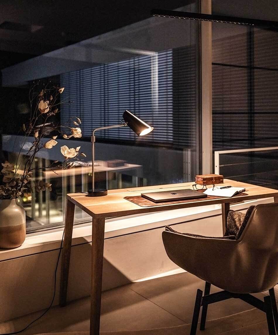 Auf einem Holzschreibtisch steht eine Birdy Tischleuchte von der Marke Northern in Schwarz / Siber. Sie beleuchtet einen Laptop sowie ein Notizbuch. Das Arbeitszimmer ist modern und minimalistisch eingerichtet.