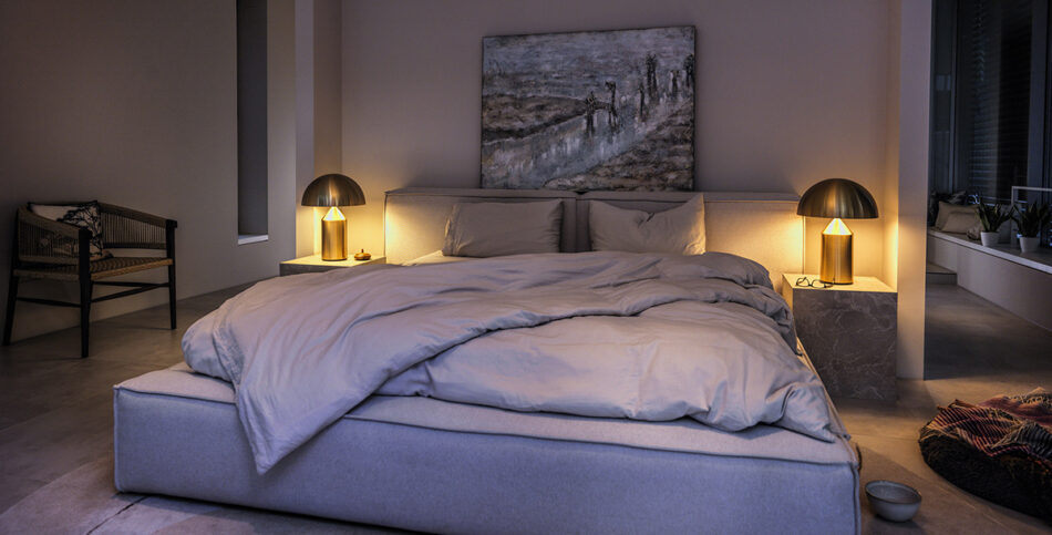 Gemütliches Schlafzimmer mit großem, grauem Bett und hellgrauer Bettdecke. Auf beiden Seiten des Bettes stehen Nachttische aus grauem Marmor, auf denen goldene Atollo Leuchten von Oluce leuchten und warmes Licht spenden. Über dem Bett hängt ein abstraktes Gemälde. Im Hintergrund links steht ein schwarzer Stuhl, rechts ist ein Fenster mit Pflanzen auf der Fensterbank. Der Raum wirkt ruhig und stilvoll eingerichtet.
