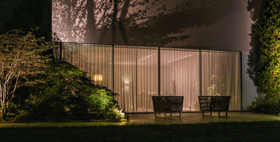 Eine von innen und außen beleuchtete Hausfassade mit Vegetationslicht sowie ein Garten mit zwei Sitzsesseln im Vordergrund.