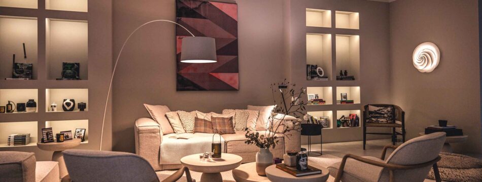 Modernes Wohnzimmer einer Villa mit unterschiedlichen Lichtquellen. Neben dem Sofa steht eine Bogenleuchte Twiggy von Foscarini. Die Wandregale werden von Lichtleisten von prediger.base beleuchtet. An der Wand hängt eine Swirl Wandleuchte von Le Klint. Das Wohnzimmer ist gemütlich eingerichtet.