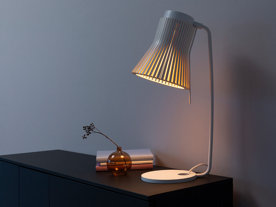 Secto Design Tischleuchte Petite 4620 auf einem dunklen Sideboard. Der charakteristische Schirm aus filigranen Holzlamellen strahlt warmes Licht nach unten ab. Neben der Leuchte stehen eine kleine Glasvase mit Zweig und einige Bücher. Die schlichte Einrichtung und die helle Wand im Hintergrund unterstreichen das skandinavische Design der Leuchte.