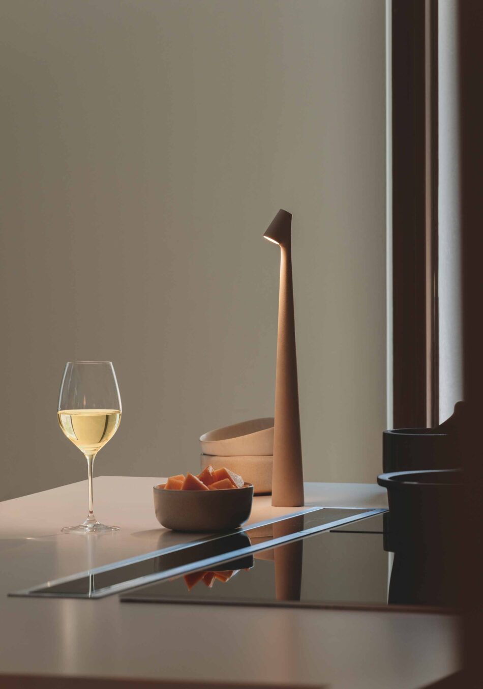 Auf einer modernen Kücheninsel steht eine Africa Tisch- und Akkuleuchte von Vibia. Sie beleuchtet ein gefülltes Weinglas und eine kleine Schale mit Obst. (Foto: Vibia)