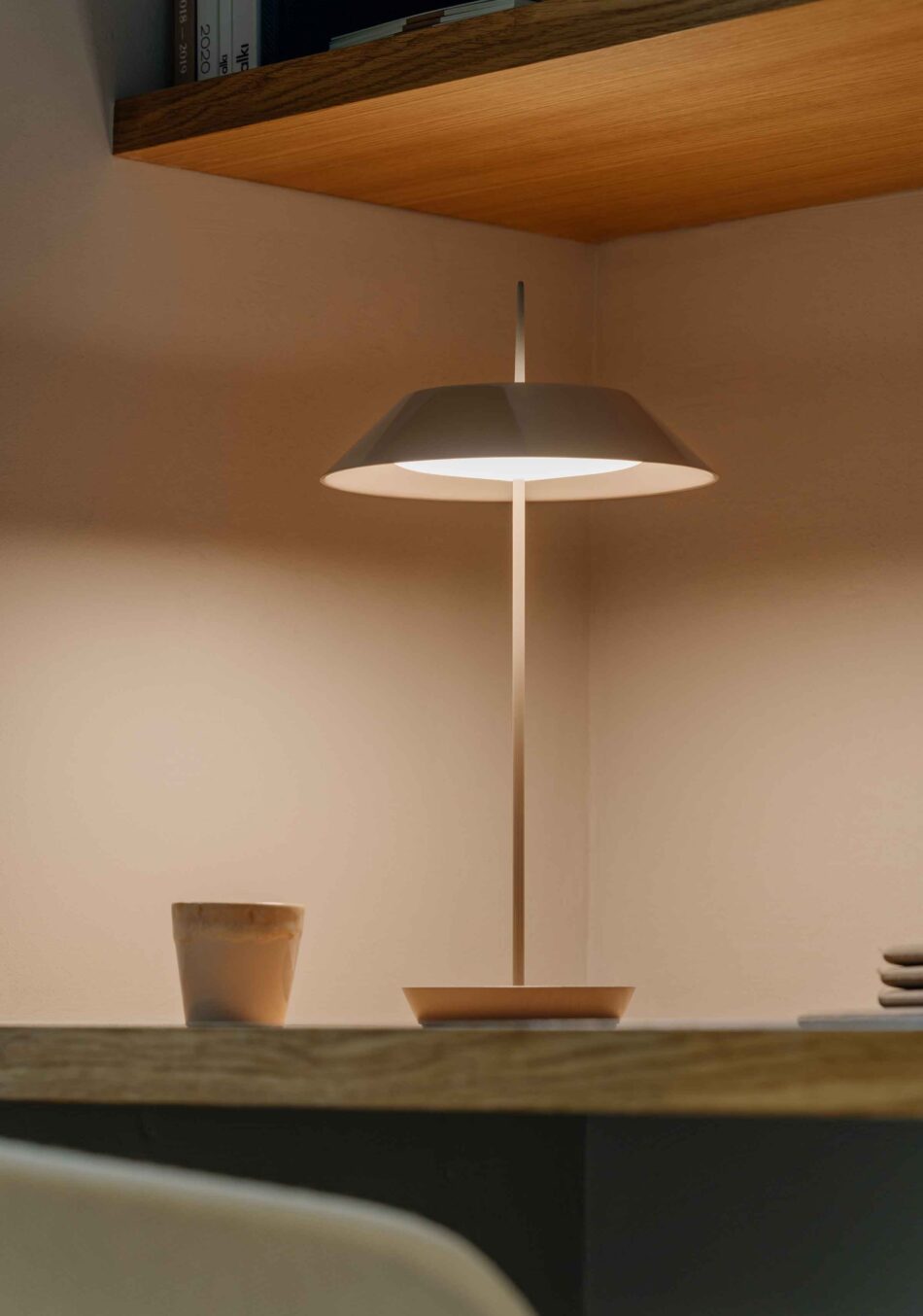 Auf einem Holzschreibtisch steht eine Mayfair Tischleuchte, die die Arbeitsfläche ausreichend beleuchtet. Über dem Schreibtisch hängt ein gefülltes Bücherregal. (Foto: Vibia)