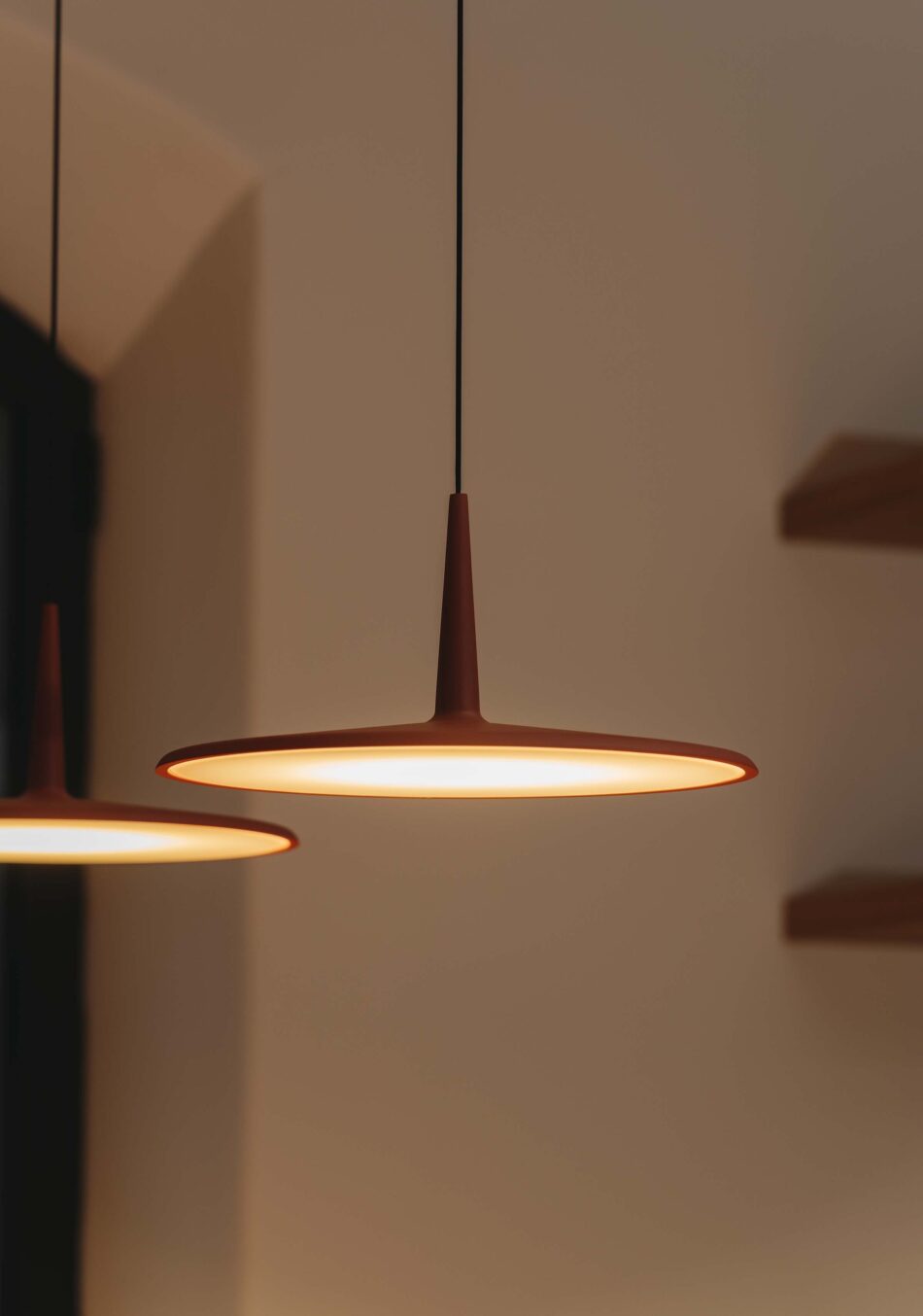 Nahaufnahme von zwei roten Skan Pendelleuchten, die einen Esstisch beleuchten. Im Hintergrund ist eine weiße Wand mit zwei Regalen zu sehen. (Foto: Vibia)