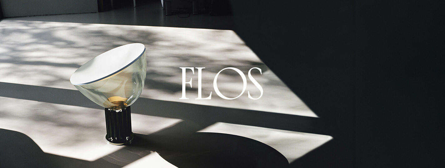 Die stilvolle Tischlampe Taccia von Flos - mit gläsernem Schirm und dunklem Sockel - steht in einem Raum mit einfallendem Sonnenlicht. Im Zentrum des Bildes ist das weiße Logo der Marke FLOS zu sehen.