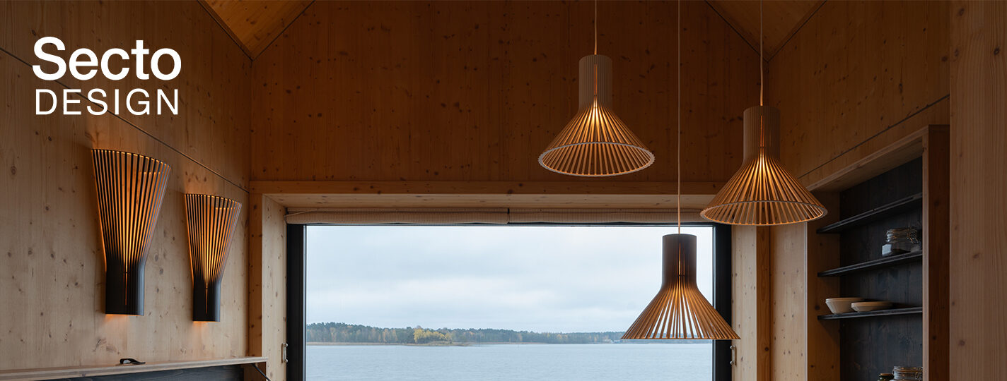 Warm beleuchteter Holzraum mit mehreren Leuchten von Secto Design. Pendel- und Wandleuchten aus Holz erzeugen ein gemütliches Lichtambiente. Im Hintergrund bietet ein großes Fenster Blick auf einen See. Links oben ist das Logo „Secto Design“ zu sehen.