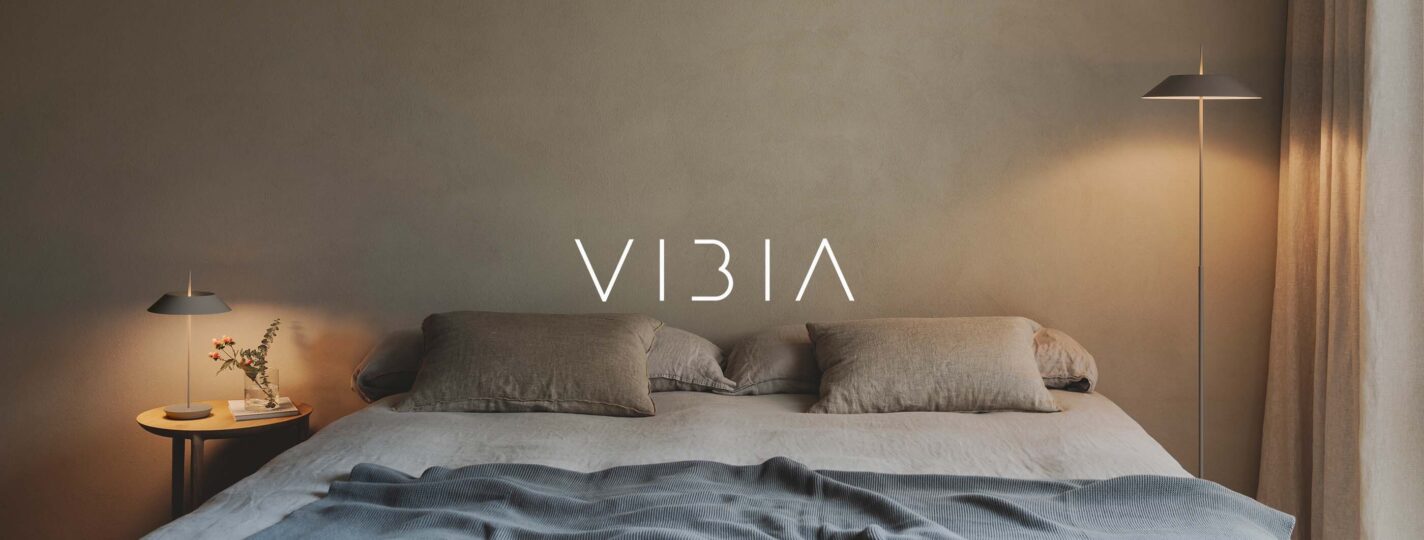 Schlichtes Schlafzimmer in Naturtönen mit einer Mayfair Tischleuchte von Vibia, die links neben dem Bett auf einem Nachttisch steht. Rechts neben dem Bett steht eine Mayfair Standleuchte von Vibia. In der Mitte des Bildes wurde das Vibia-Logo mit weißen Schriftzug platziert. (Foto: Vibia)