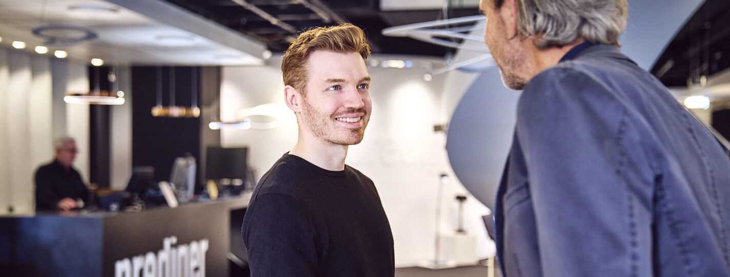 Ein junger Lichtberater lächelt und spricht mit einem Kunden im Prediger Showroom. Im Hintergrund ist der Empfangsbereich von Prediger zu sehen. (Foto: Johannes Arlt)