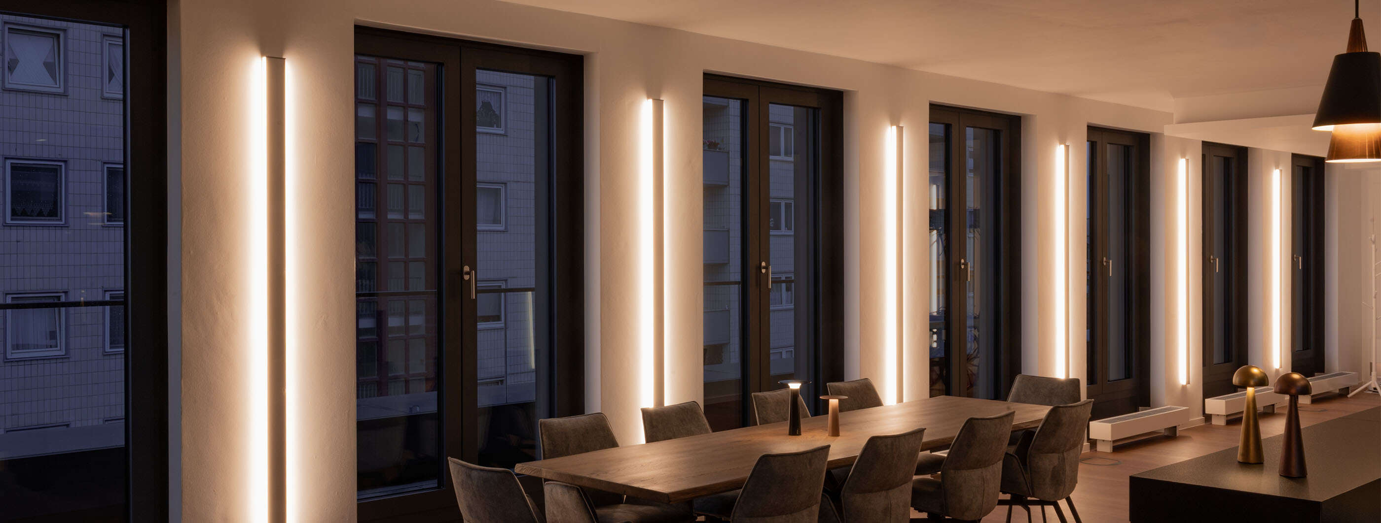 Großer Konferenzraum mit LED-Wandprofilen von Sigor und zwei Lix Akkuleuchten von IP44.DE, die auf dem langen Konfernztisch stehen. Rechts auf dem Tresen stehen zwei weitere p.141 Akkuleuchten von prediger.base.