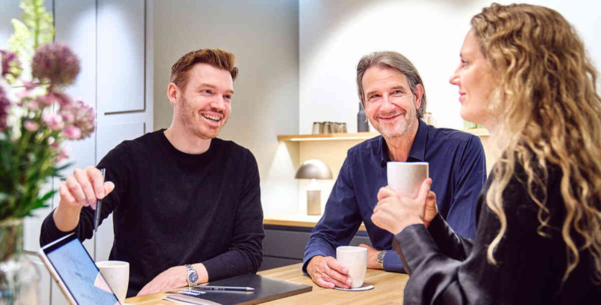 Gespräch zwischen Kunden und Lichtberater, die an einem Küchentisch sitzen und eine Lichtplanung auf dem Laptop besprechen. Die Kunden halten Kaffeebecher in der Hand. Im Hintergrund ist eine moderne Küche mit einer kleinen Atollo Tischleuchte von Oluce auf dem Küchentresen zu sehen.