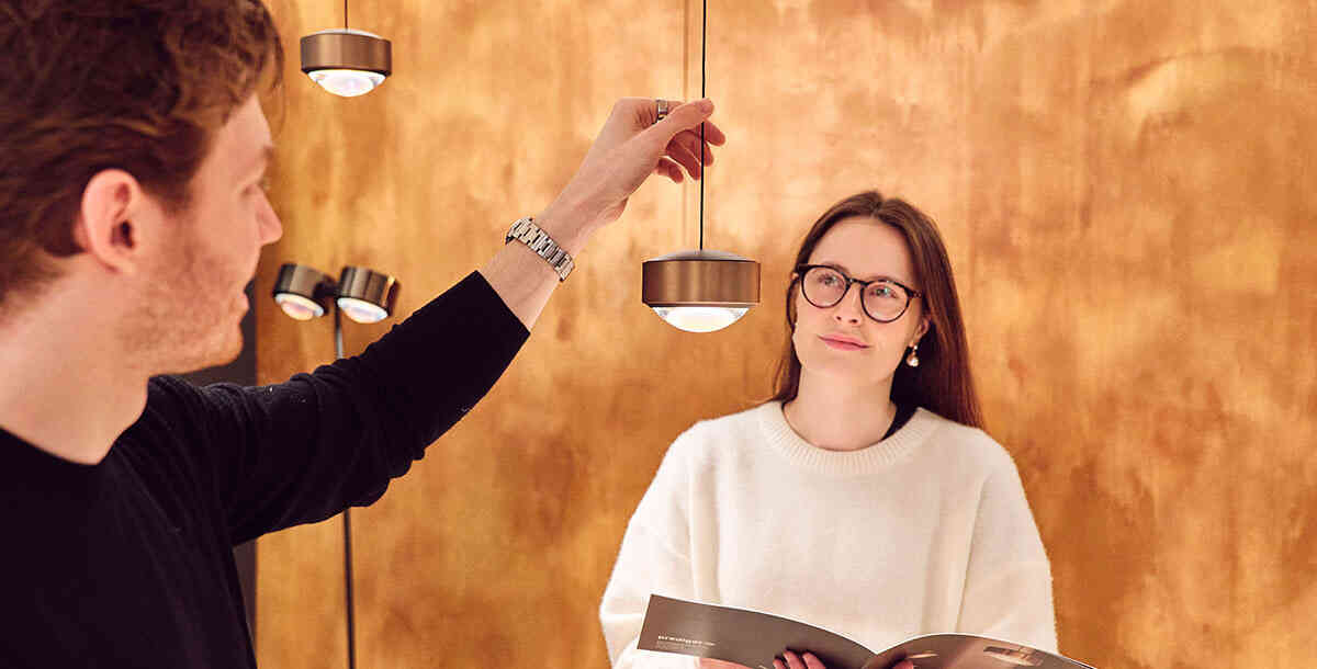 Ein Lichtberater stellt einer Kundin die s.100 Pendelleuchte von prediger.select vor. Die Kundin hat dabei den prediger.select Katalog in der Hand und hört aufmerksam zu.