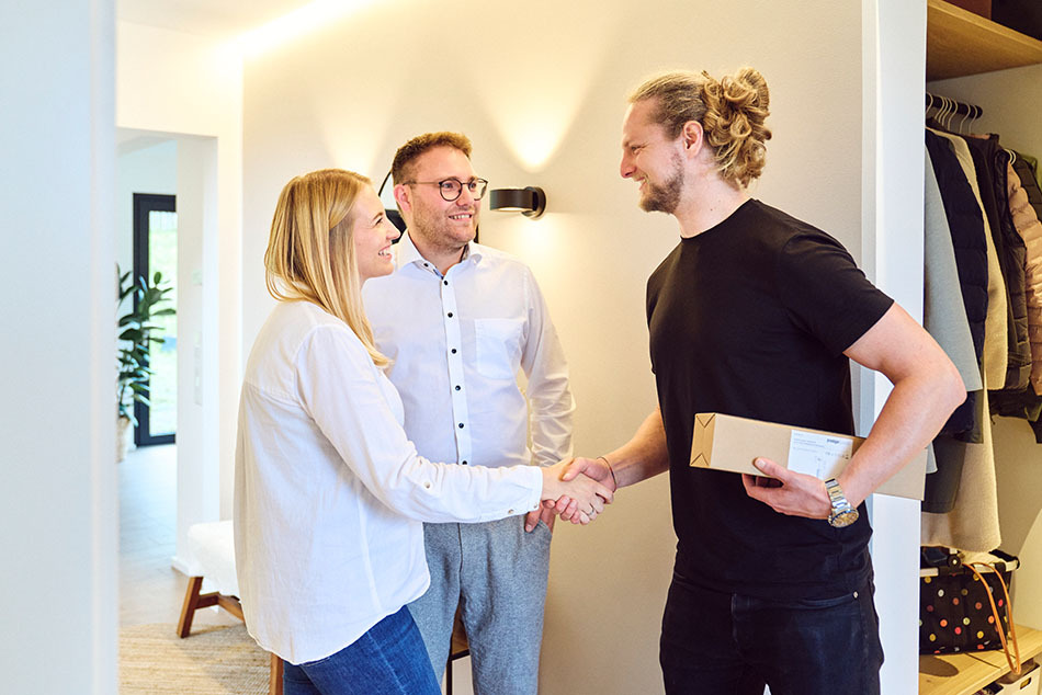 Ein Lichtberater überreicht im Zuhause der Kunden ein Paket und begrüßt sie mit einem Handschlag. Die beiden Kunden stehen ihm freundlich gegenüber.