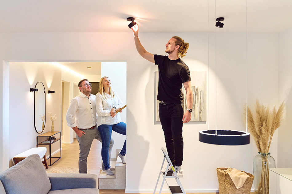 Lichtberater von Prediger richtet eine schwarze p.011 Deckenleuchte im Wohnzimmer aus während er auf einem Tritt steht. Die beiden Kunden schauen aufmerksam zu.