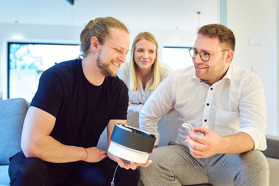 Lichtberater von Prediger stellt einem Kunden eine Leuchte von CHRISTOPH vor. Beide Personen sitzen auf einem grauen Sofa im Wohnzimmer des Kunden.